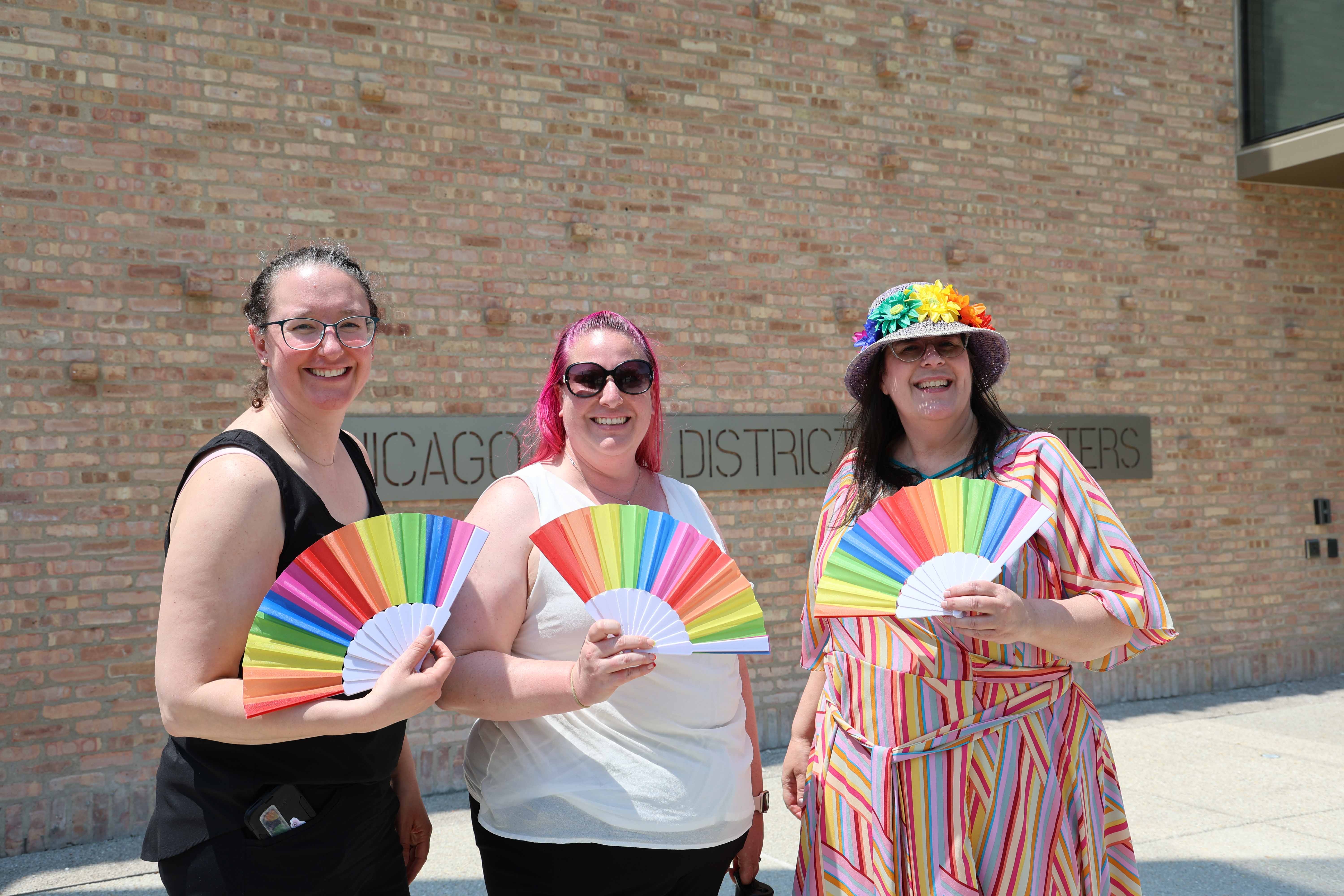 Three smiling people hold rainbow fans in front of a brick wall.